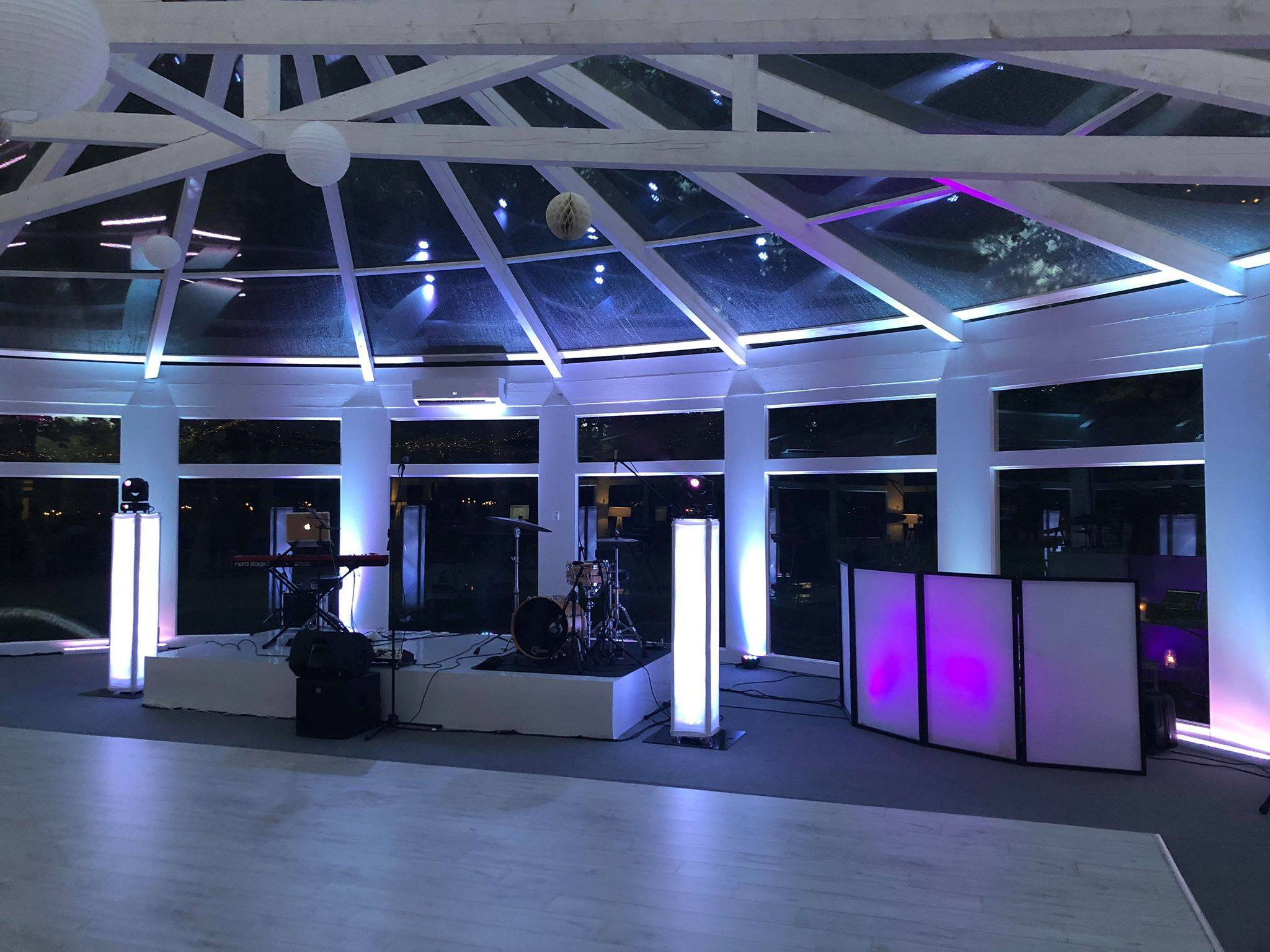 Scena z instrumentami muzycznymi i stanowiskiem DJ-a oświetlona białymi i fioletowymi światłami w przeszklonej sali z drewnianą konstrukcją dachu i lampionami.