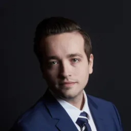 Portret młodego mężczyzny w granatowym garniturze i krawacie w paski na ciemnym tle, z delikatnym uśmiechem i zaczesanymi do tyłu włosami.