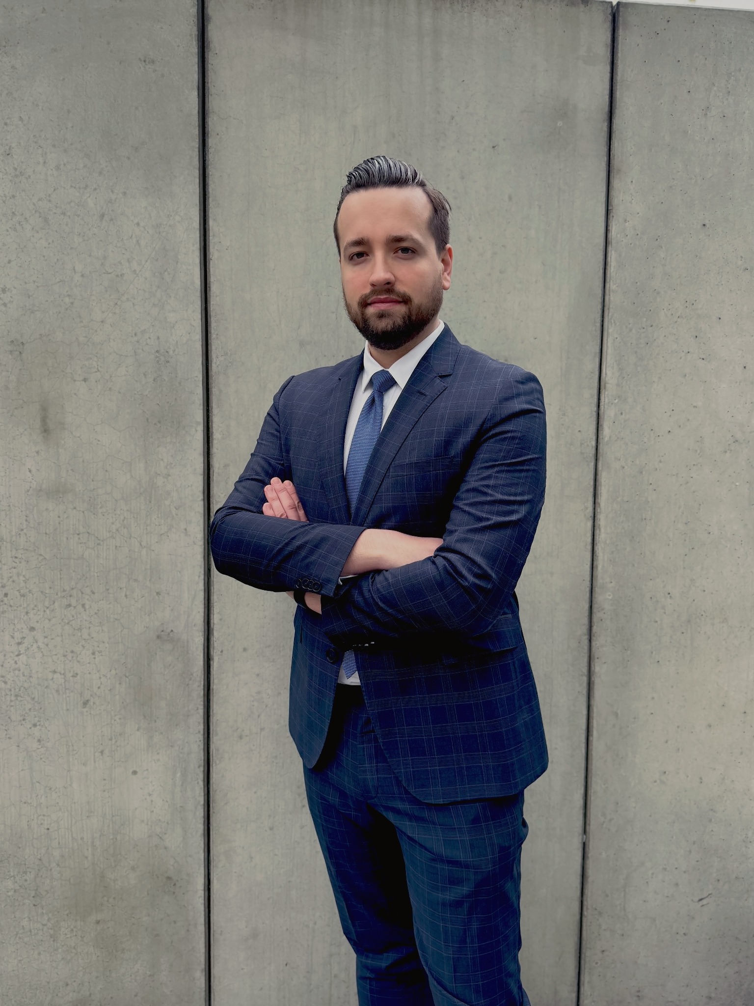 Mężczyzna w granatowym garniturze w kratę z założonymi rękami na tle betonowej ściany z widocznymi łączeniami.