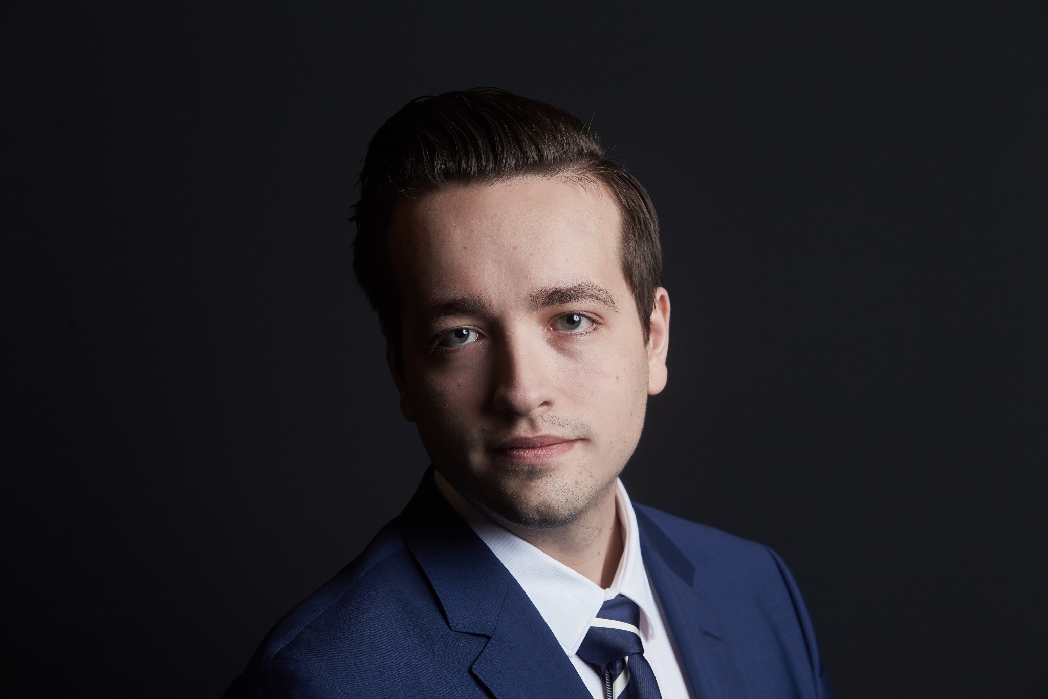 Portret młodego mężczyzny w granatowym garniturze i krawacie w paski na ciemnym tle, z delikatnym uśmiechem i zaczesanymi do tyłu włosami.