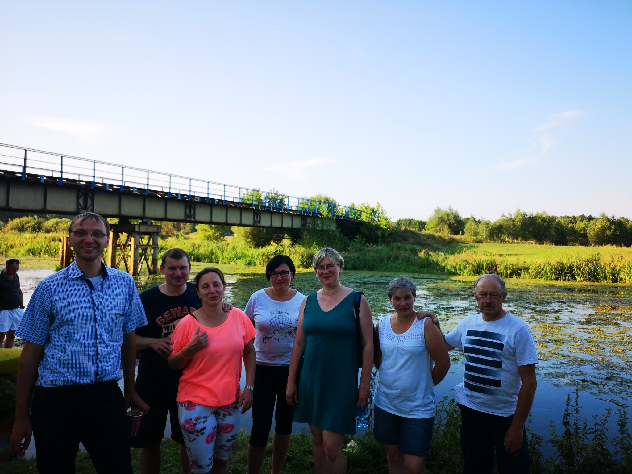 Grupa ośmiu osób stoi na tle mostu kolejowego i zarośniętego jeziora podczas integracji firmowej, mężczyzna w niebieskiej koszuli trzyma kubek.