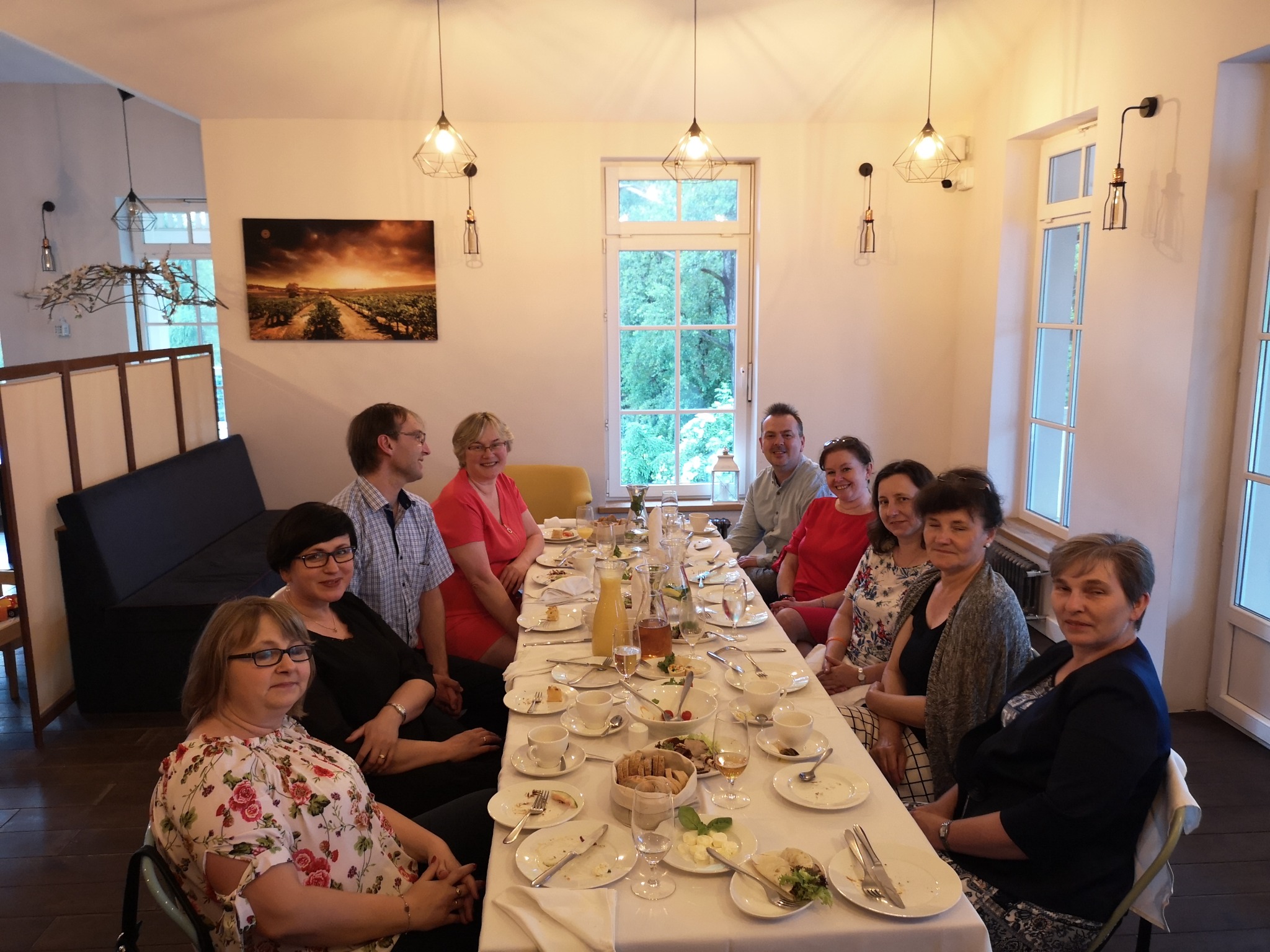 Grupa osób siedzi przy nakrytym stole z resztkami jedzenia, w tle obraz z winnicą i nowoczesne lampy.