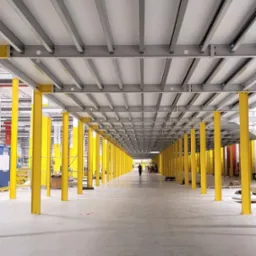 Wnętrze hali przemysłowej w trakcie budowy, widoczne żółte stalowe słupy podtrzymujące szarą konstrukcję stropu, w tle sylwetka osoby.