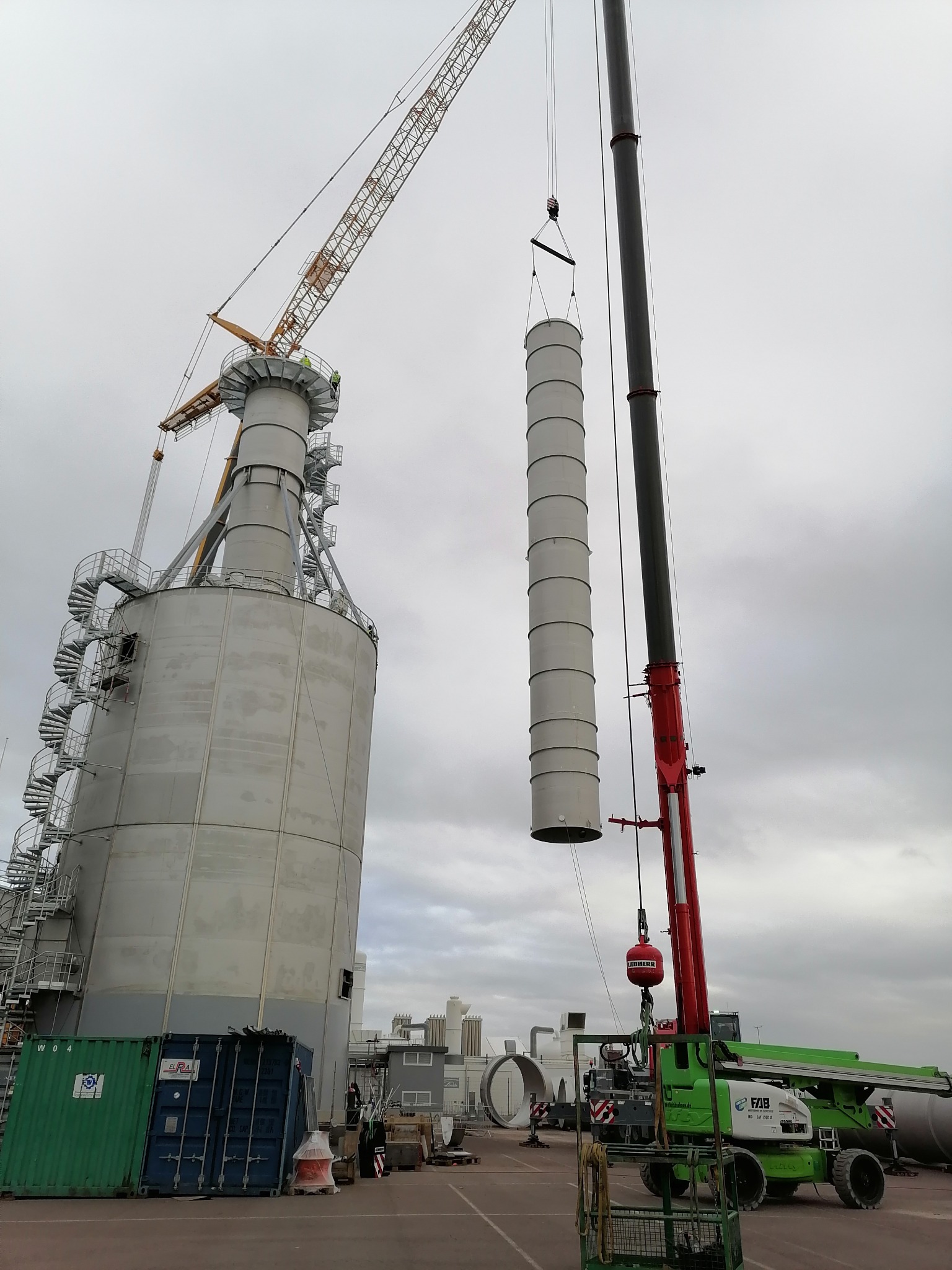 Montaż cylindrycznego elementu konstrukcji stalowej za pomocą dwóch dźwigów na placu budowy, widoczne kontenery i podnośnik koszowy.