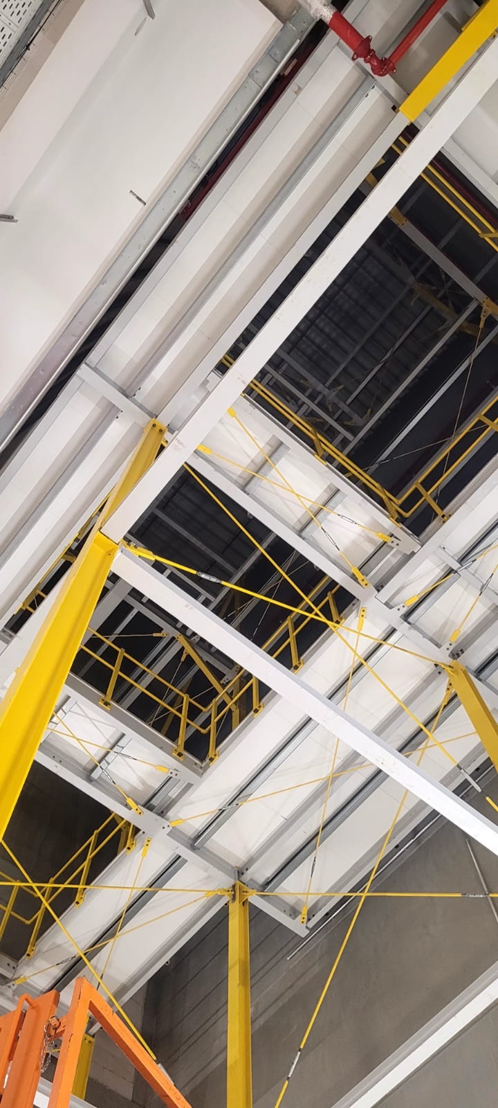 Wnętrze hali w trakcie budowy z widoczną konstrukcją stalową, żółtymi słupami nośnymi i elementami mocującymi pod sufitem, sfotografowane z dołu pod kątem.