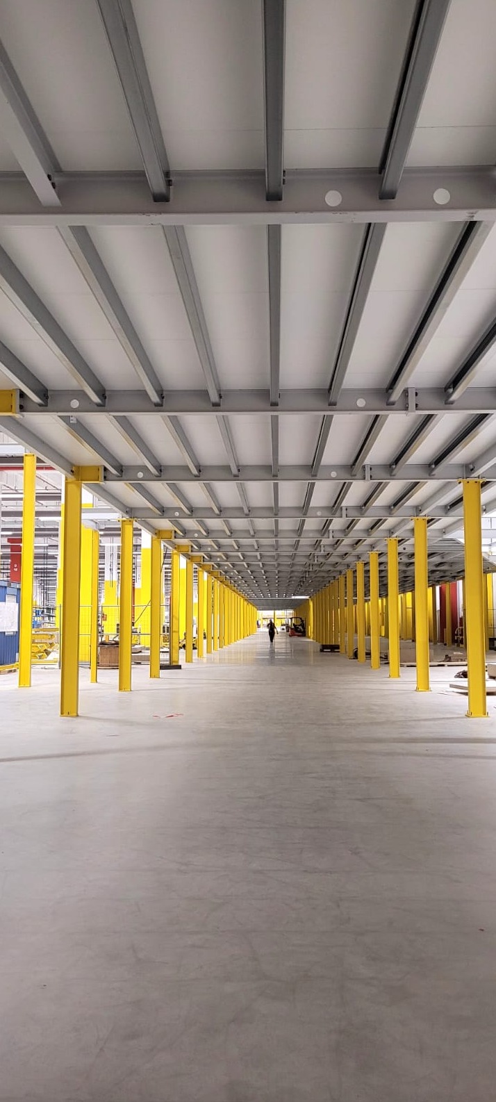 Wnętrze hali przemysłowej w trakcie budowy, widoczne żółte stalowe słupy podtrzymujące szarą konstrukcję stropu, w tle sylwetka osoby.
