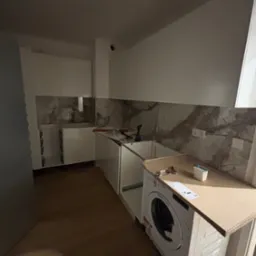 Montaż zabudowy kuchennej: białe szafki z marmurowym panelem, pralka wkomponowana w zabudowę, narzędzia na blacie. Widok na wnętrze w trakcie wykończenia.