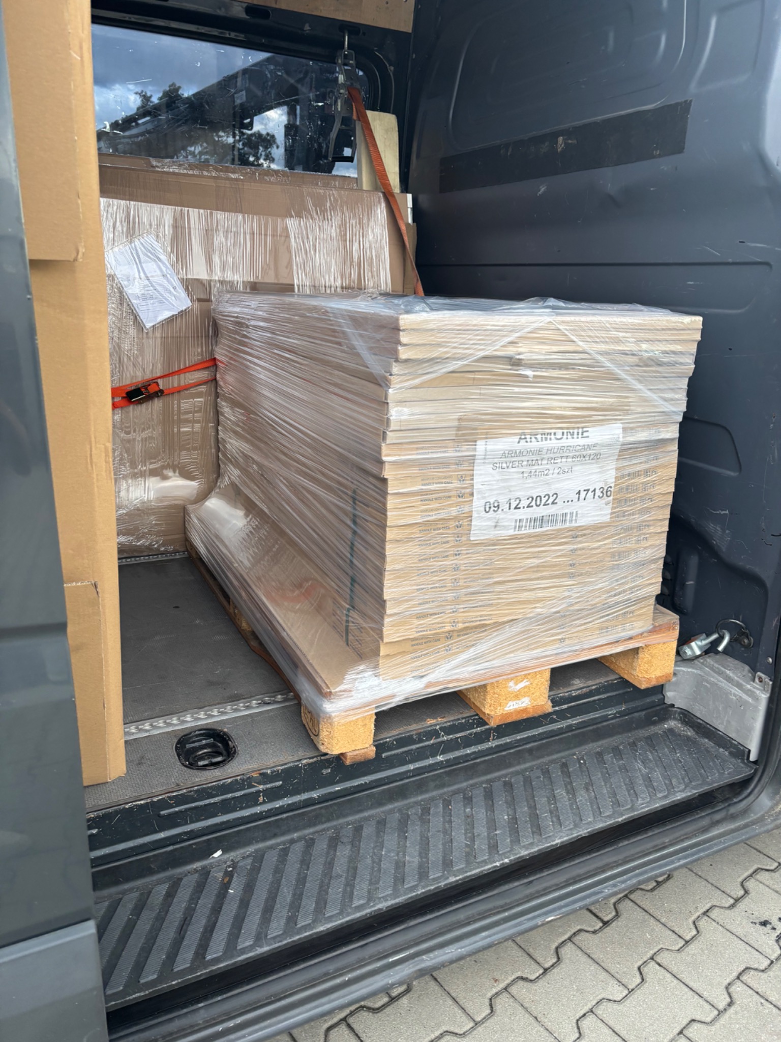 Paleta z płytkami ceramicznymi, zabezpieczona folią, w przestrzeni ładunkowej busa. Widoczna etykieta z nazwą 'Armonie Hurricane Silver Mat Rett 60x120'.