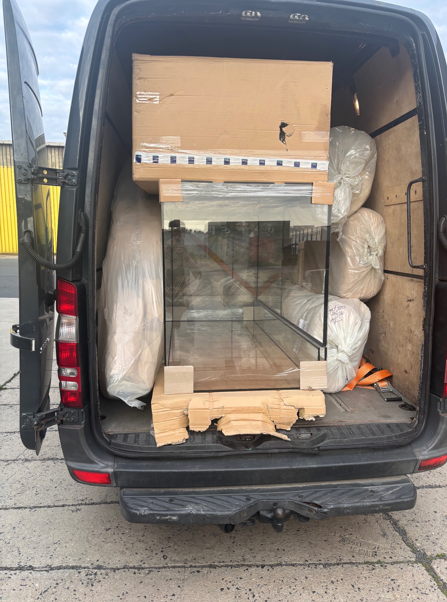 Transport akwarium i worków z materiałami w bagażniku busa. Widoczne kartony, drewniane elementy zabezpieczające i pasy mocujące ładunek. Otwarte drzwi pojazdu.
