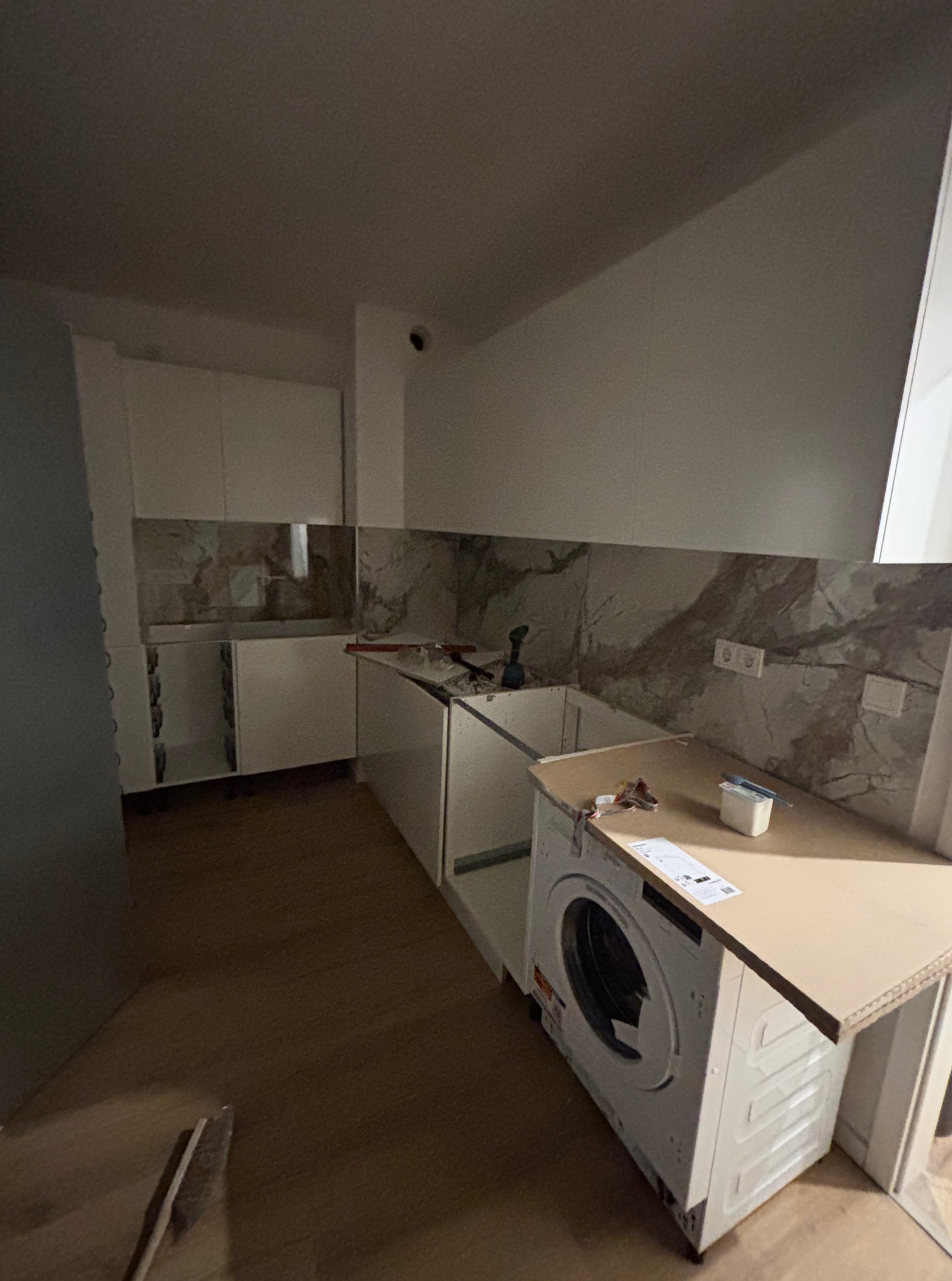 Montaż zabudowy kuchennej: białe szafki z marmurowym panelem, pralka wkomponowana w zabudowę, narzędzia na blacie. Widok na wnętrze w trakcie wykończenia.