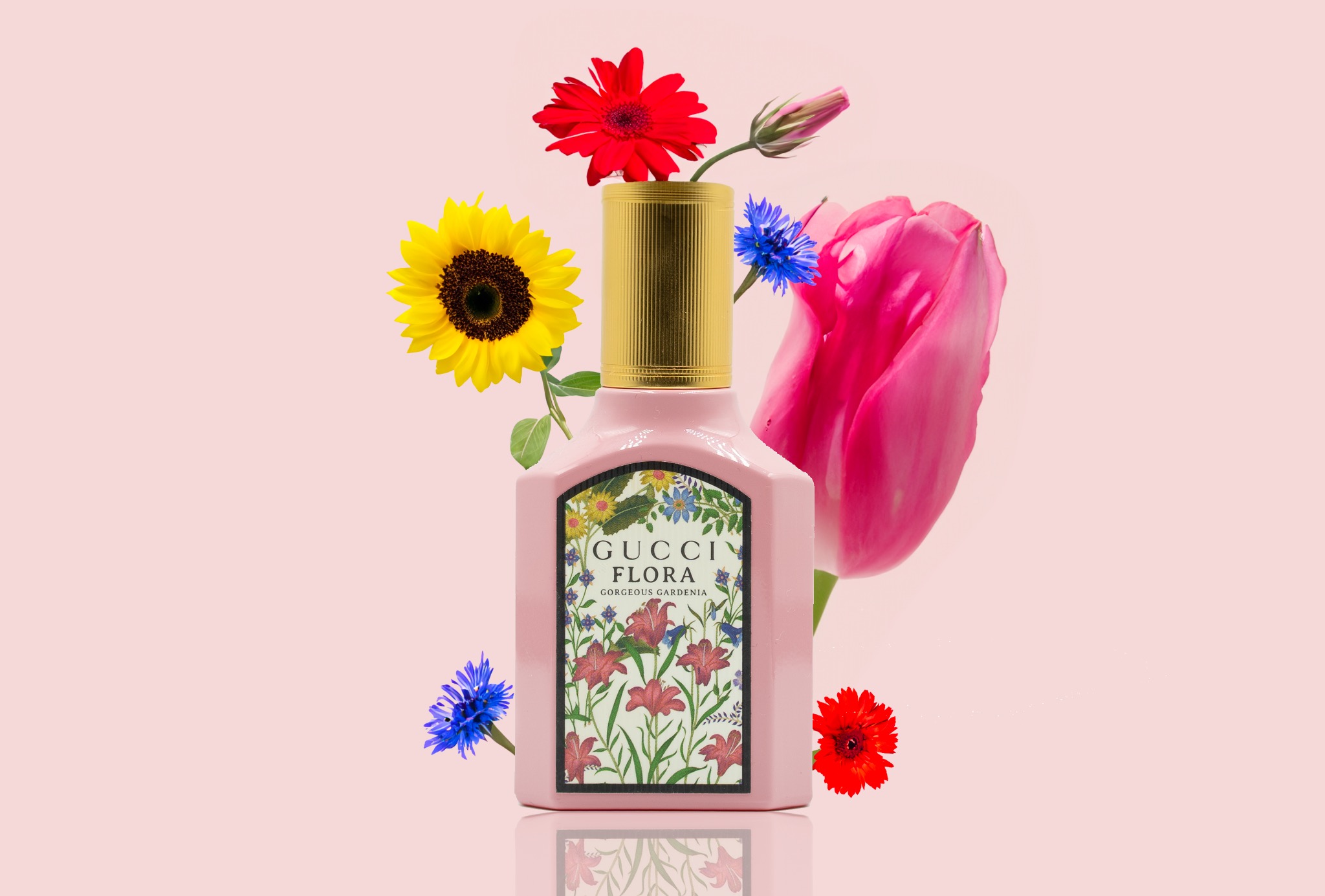 Różowy flakon perfum Gucci Flora Gorgeous Gardenia otoczony kwiatami: słonecznikiem, gerberą, chabrem i tulipanem na jasnoróżowym tle.
