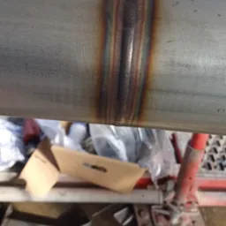 Spoina na rurze ze stali nierdzewnej, widoczne przebarwienia termiczne, fragment rusztowania w tle.