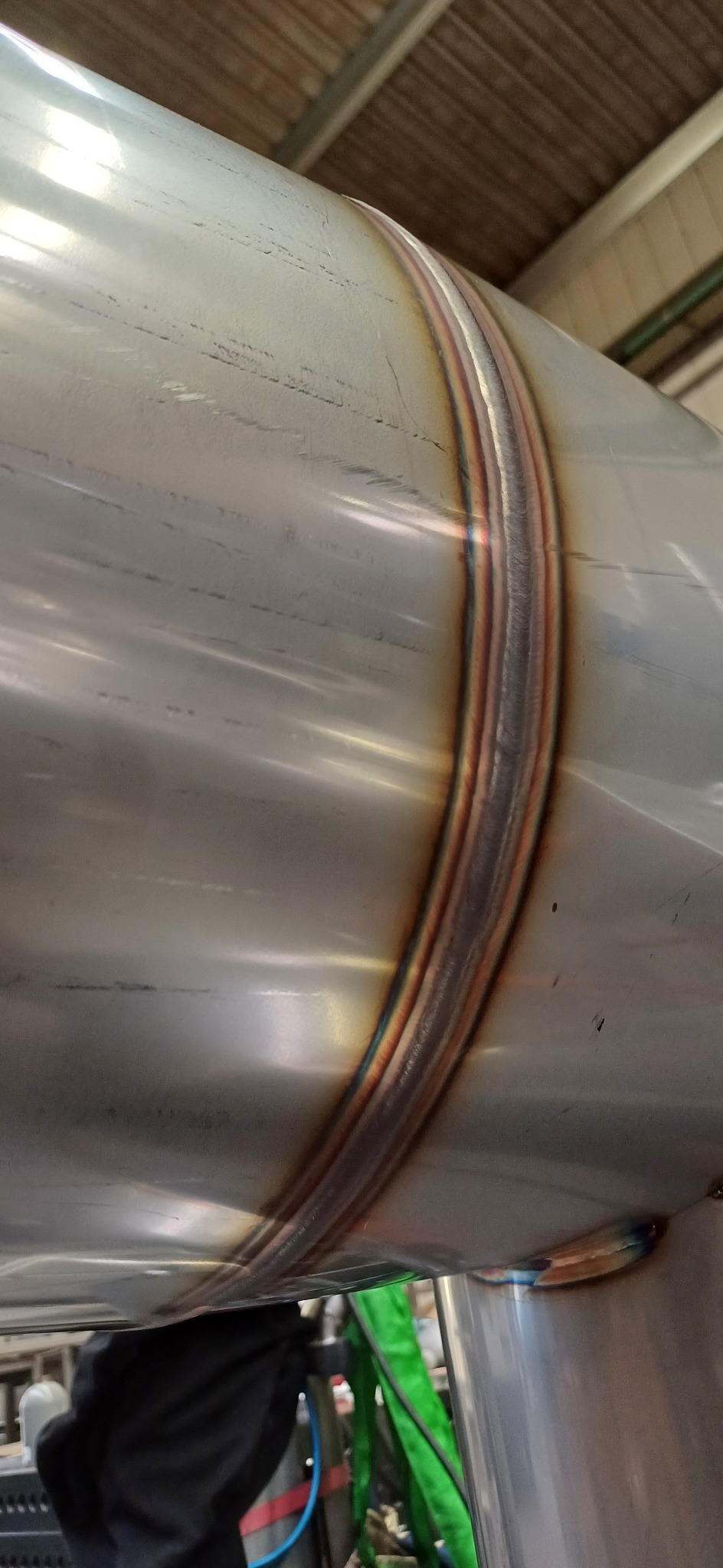 Spoina obwodowa na rurze ze stali nierdzewnej, widoczne przebarwienia termiczne w kolorach tęczy, otoczenie warsztatowe.