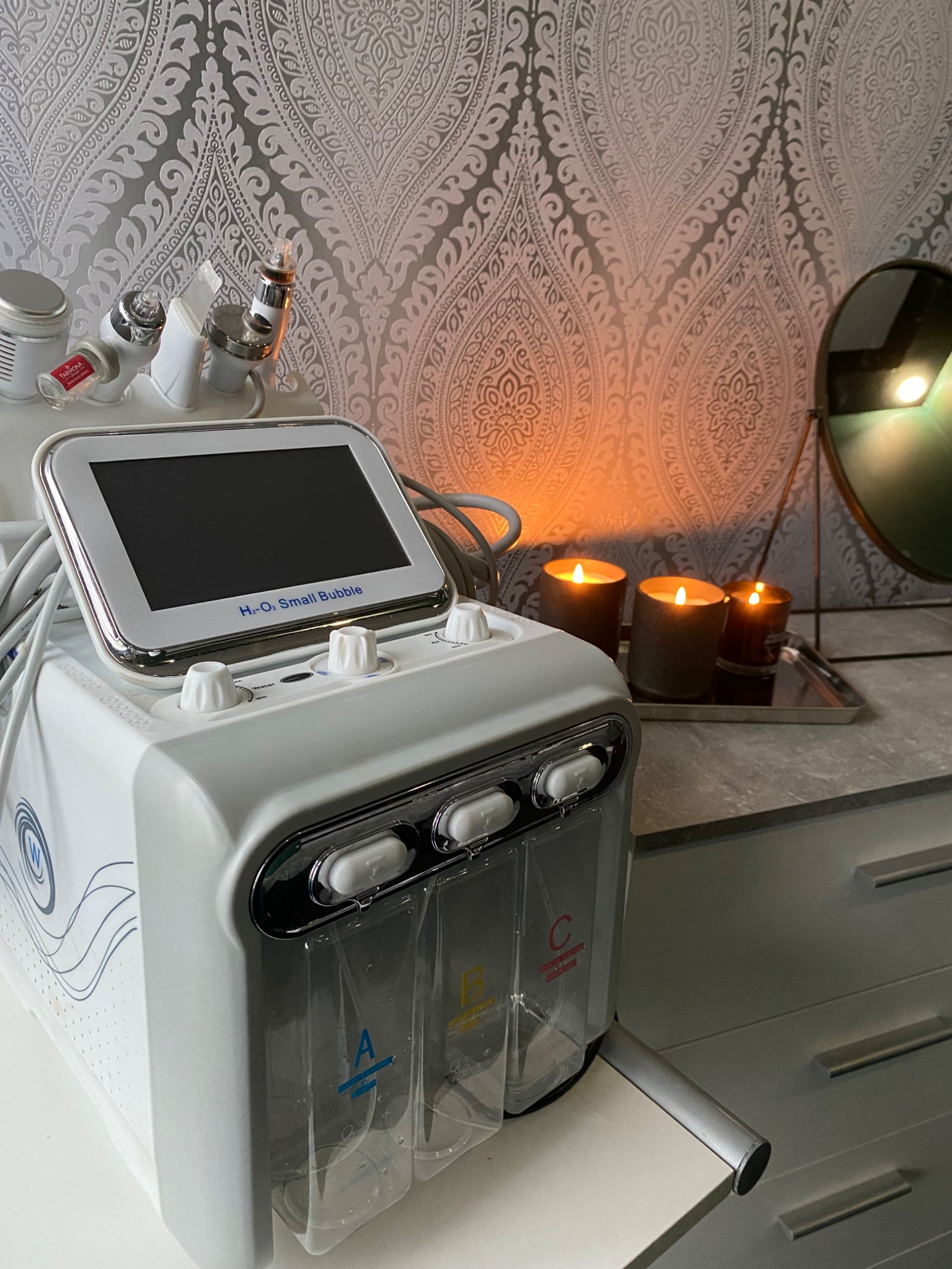 Aparat do hydrabrazji z wyświetlaczem i pojemnikami na płyny A, B, C, ustawiony na blacie w gabinecie kosmetycznym, w tle ozdobna tapeta i zapalone świece.