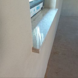 Duomit Pro - Świeżo nałożony, jasnoszary tynk maszynowy na ścianie wewnętrznej z widocznym parapetem okiennym z połyskiem i surową posadzką w pomieszczeniu w trakcie remontu.