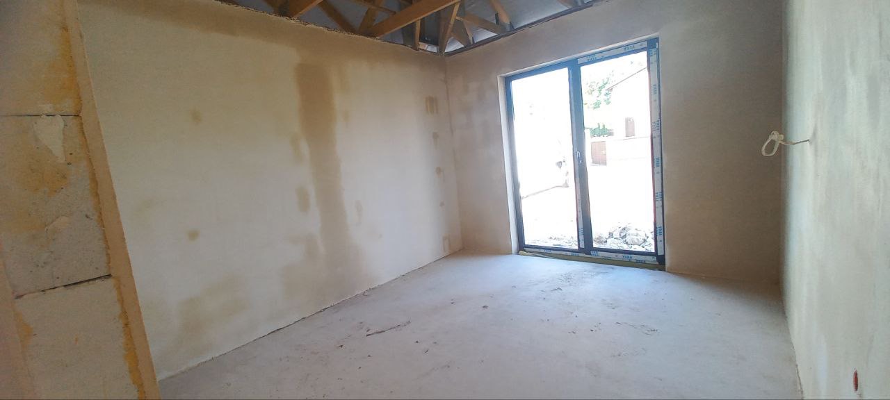 Pokój w stanie surowym po nałożeniu warstwy tynku; widoczne ślady wilgoci na ścianach, drewniana konstrukcja więźby dachowej pod sufitem oraz duże okno balkonowe.