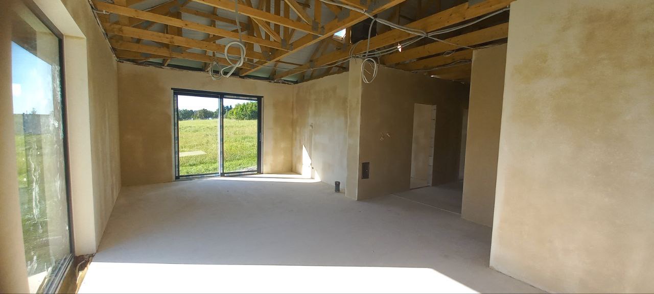 Wnętrze domu w trakcie prac wykończeniowych, ściany pokryte świeżym tynkiem w odcieniach beżu, surowa drewniana konstrukcja dachu z widocznymi kablami, duże okno przesuwne z widokiem na zielone pole.