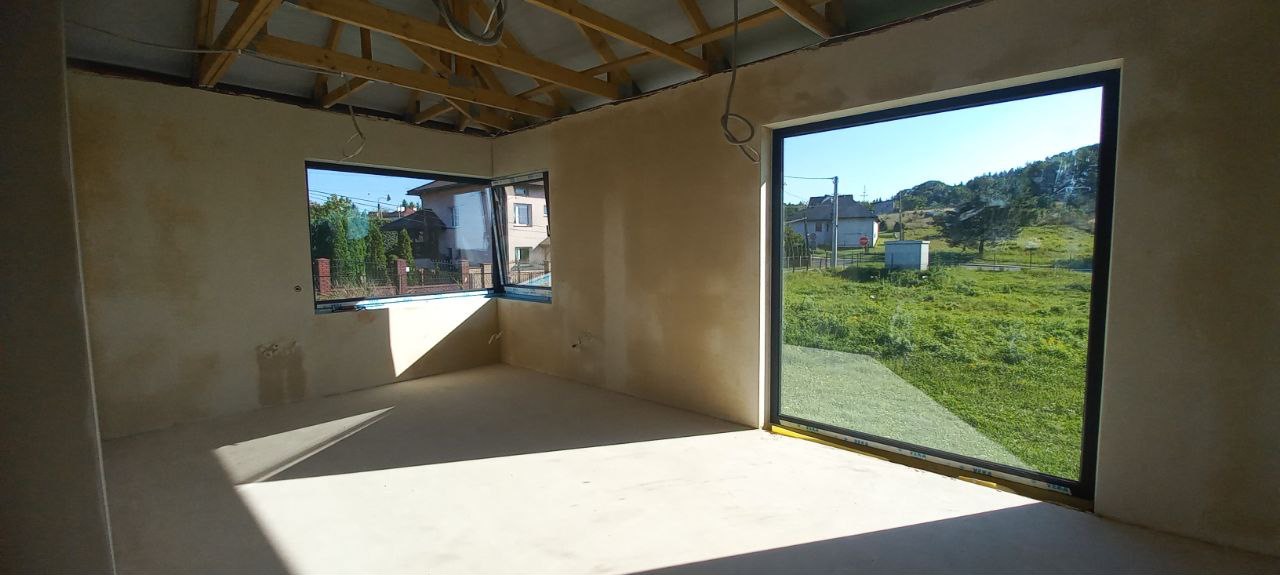 Surowe ściany wewnętrzne z oknami w nowym budynku, widoczne drewniane krokwie stropu, przygotowane do dalszych prac wykończeniowych, z widokiem na zewnątrz przez okna.