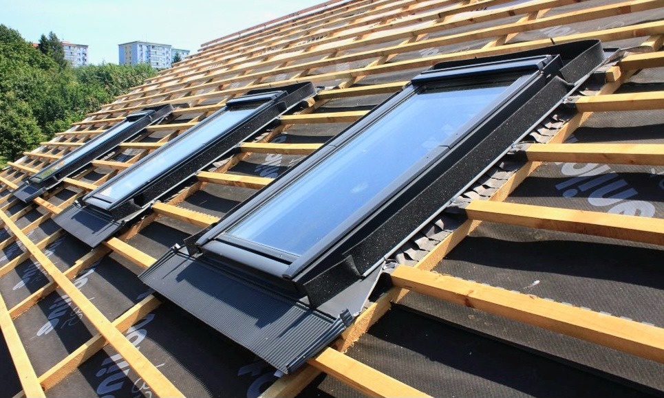 Montaż trzech okien dachowych na przygotowanej konstrukcji z łat i kontrłat, widoczna membrana dachowa, w tle budynki mieszkalne i drzewa.