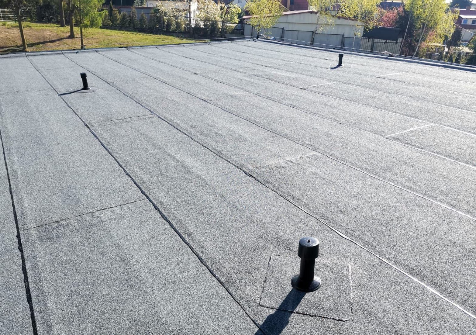 Szary dach płaski pokryty papą termozgrzewalną z widocznymi kominkami wentylacyjnymi, na tle zieleni i zabudowań.