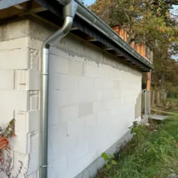 Nowa, błyszcząca, metalowa rynna pionowa i pozioma zamontowana na ścianie budynku z białych bloczków betonowych, z widocznymi elementami dachu i drzewami w tle.