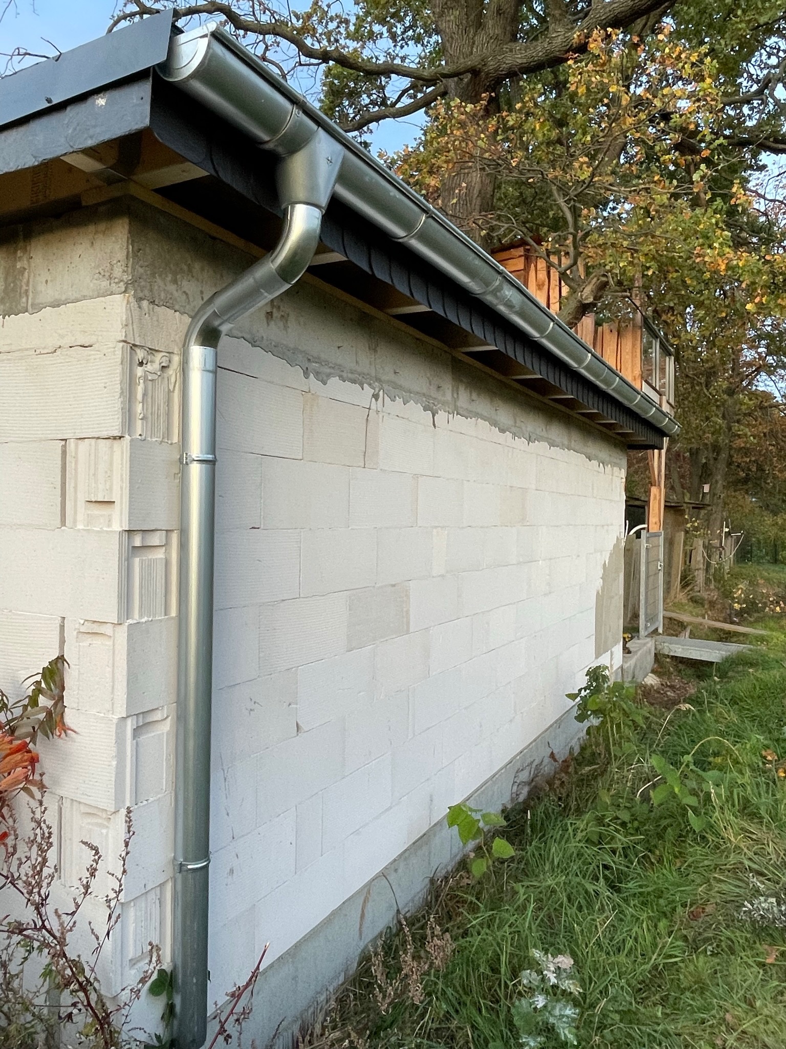 Nowa, błyszcząca, metalowa rynna pionowa i pozioma zamontowana na ścianie budynku z białych bloczków betonowych, z widocznymi elementami dachu i drzewami w tle.