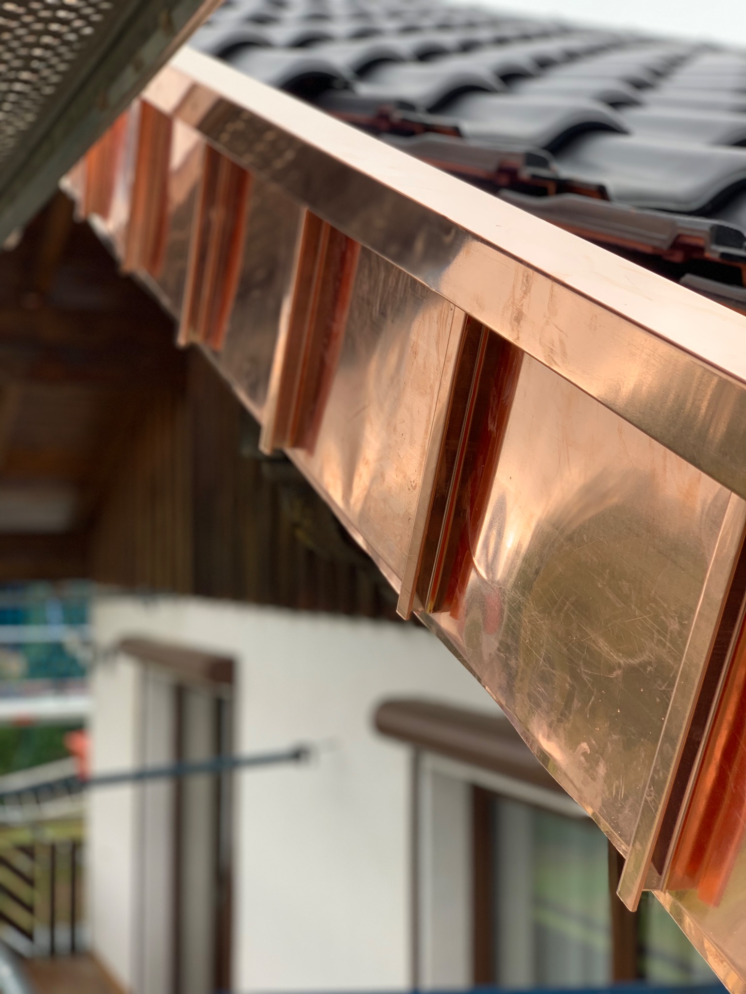 Miedziana rynna dachowa z bliska, widoczne łączenia i fragment dachu z dachówką ceramiczną oraz fragment elewacji budynku w tle.