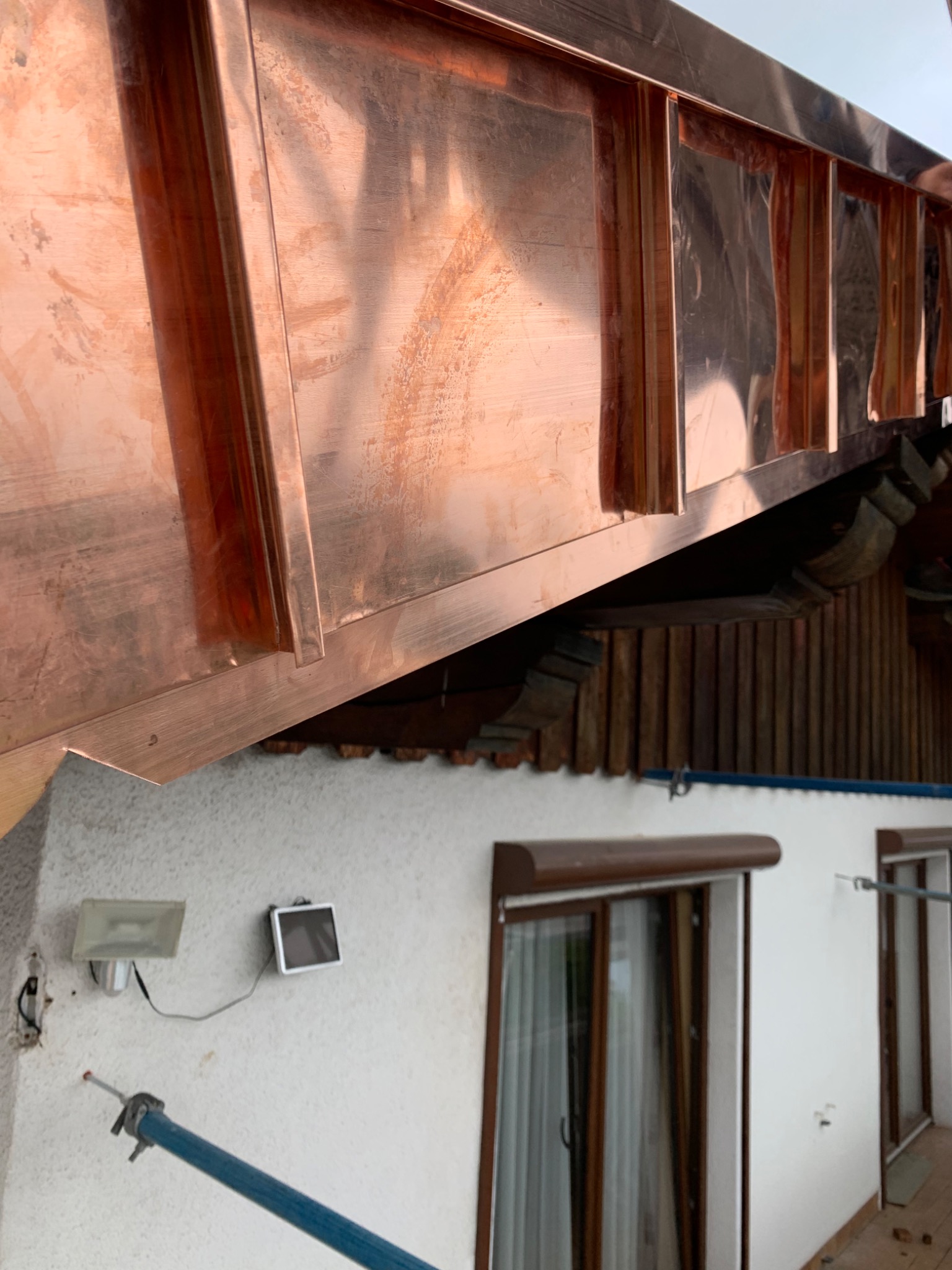 Miedziana obróbka blacharska dachu, widoczne elementy mocowania i fragment elewacji z oknami.