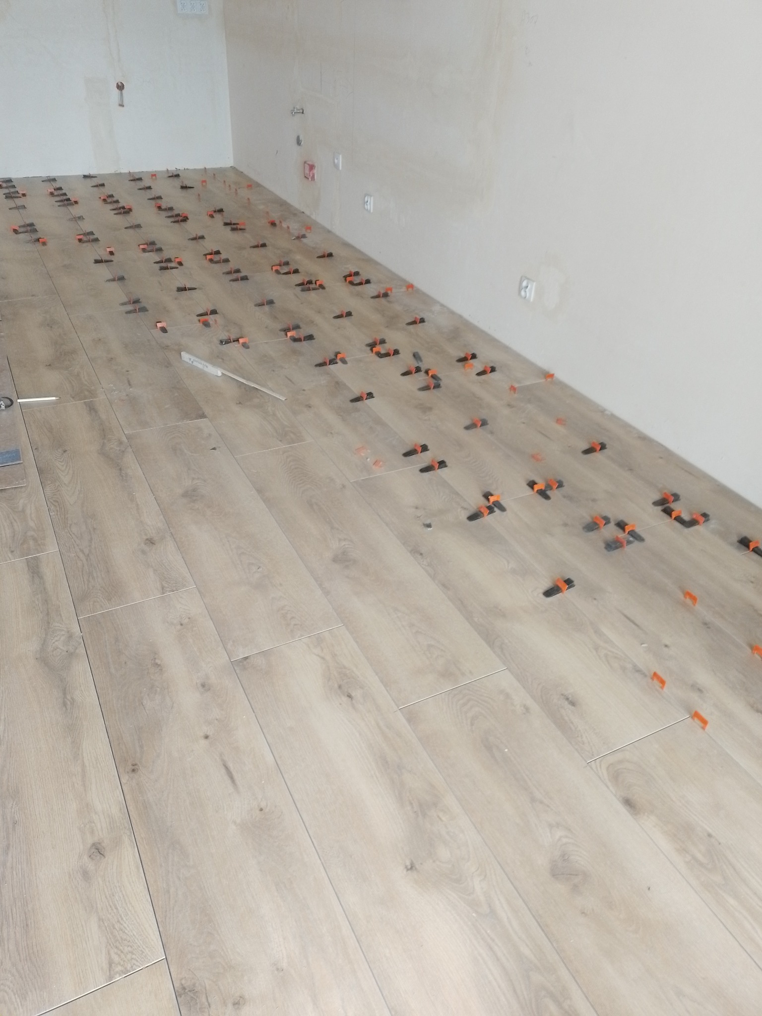 Układanie podłogi z płytek imitujących drewno z użyciem klipsów poziomujących w jasnym pomieszczeniu, widoczne narzędzia glazurnicze.