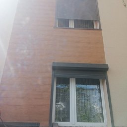 Przedsiębiorstwo wielobranżowe - Elewacja budynku z drewnopodobną okładziną, widoczne dwa okna z roletami zewnętrznymi, jedno z roletą opuszczoną, drugie podniesioną, fragment dachu i roślinności.