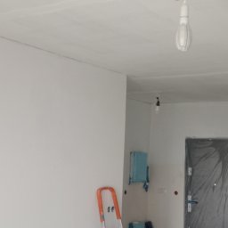 Przedsiębiorstwo wielobranżowe - Świeżo pomalowane białe ściany i sufit w trakcie prac wykończeniowych, drabina oparta o ścianę, zabezpieczone folią drzwi w tle.