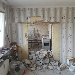 Przedsiębiorstwo wielobranżowe - Demontaż ściany działowej między pokojem a kuchnią, widoczne gruz, odsłonięte instalacje, drabina i kuchenka w tle, ściany z tapetą w pokoju.