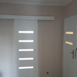 Białe, przesuwne drzwi z poziomymi przeszkleniami, zamontowane w jasnym wnętrzu. Widoczna r&oacute;wnież standardowa biała ościeżnica z klamką.