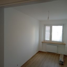 Przedsiębiorstwo wielobranżowe - Jasne, pomalowane na biało wnętrze pokoju z drewnianą podłogą, widoczne przez framugę drzwi. Pod oknem grzejnik i stos paneli podłogowych.