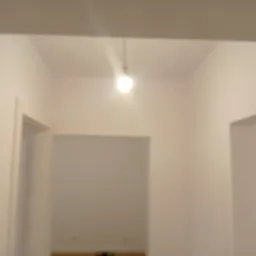 Świeżo pomalowane, jasne ściany wewnątrz pomieszczenia z widocznymi otworami drzwiowymi. Centralnie umieszczona żarówka oświetla wnętrze. Minimalistyczny wystrój.