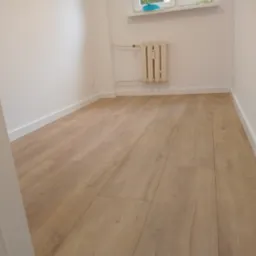 Jasne panele podłogowe w pokoju z białymi ścianami i grzejnikiem. Widok zza framugi drzwi, neutralne oświetlenie, minimalistyczny wystrój.