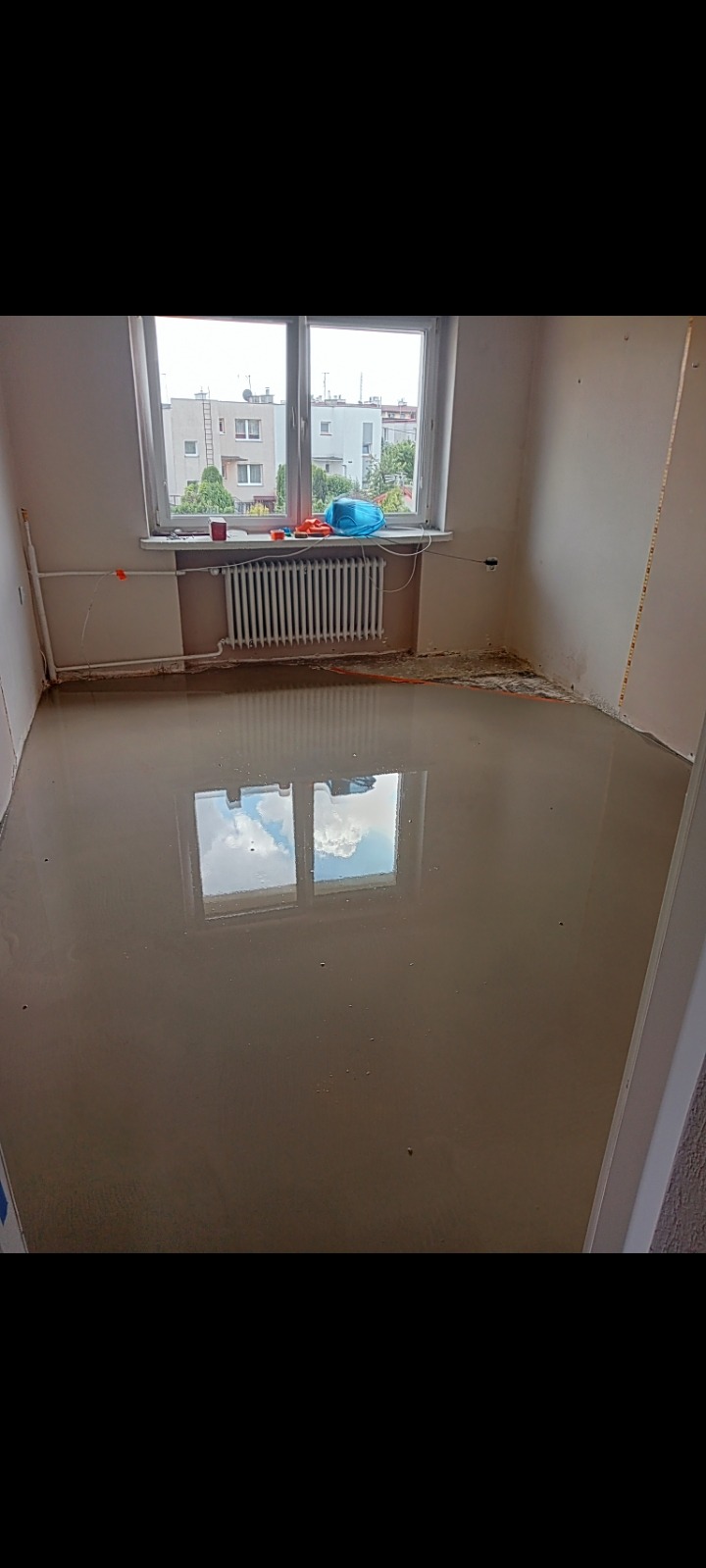 Wylany samopoziomujący podkład podłogowy w pokoju, odbicie okna w mokrej powierzchni, grzejnik pod oknem, narzędzia i niebieska torba na parapecie.