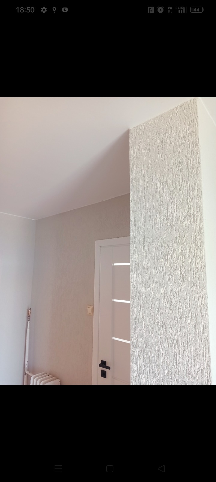 Fragment pokoju po nałożeniu strukturalnej, jasnej gładzi na ścianę, widoczna biała ościeżnica z czarną klamką i fragment grzejnika.