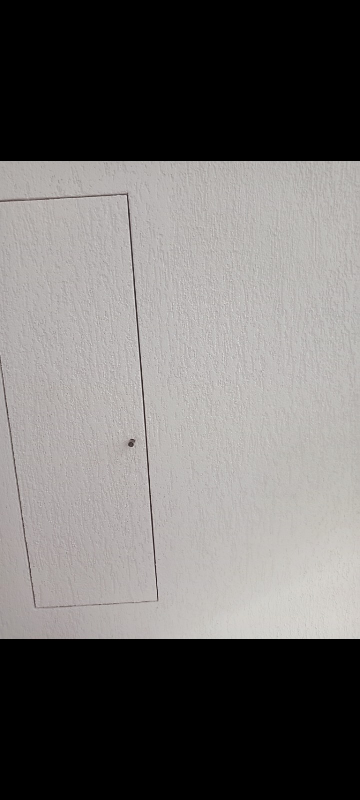 Biała klapa rewizyjna w ścianie z szarą, chropowatą strukturą, zabezpieczona małym metalowym zatrzaskiem.