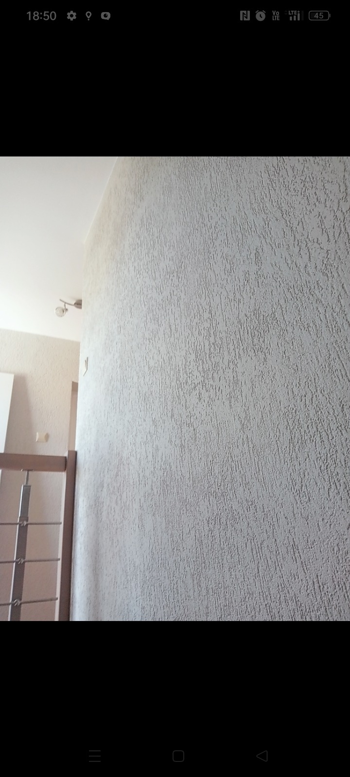 Ściana wewnętrzna z fakturą baranka w jasnym odcieniu, widoczny fragment sufitu i oświetlenia, w tle fragment klatki schodowej.