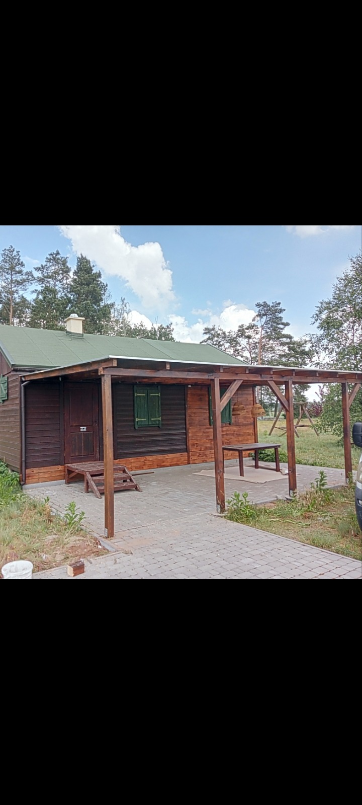 Drewniany domek z zadaszeniem tarasowym, pomalowany na ciemny brąz, z zielonymi okiennicami i schodami wejściowymi, otoczony zielenią i kostką brukową.