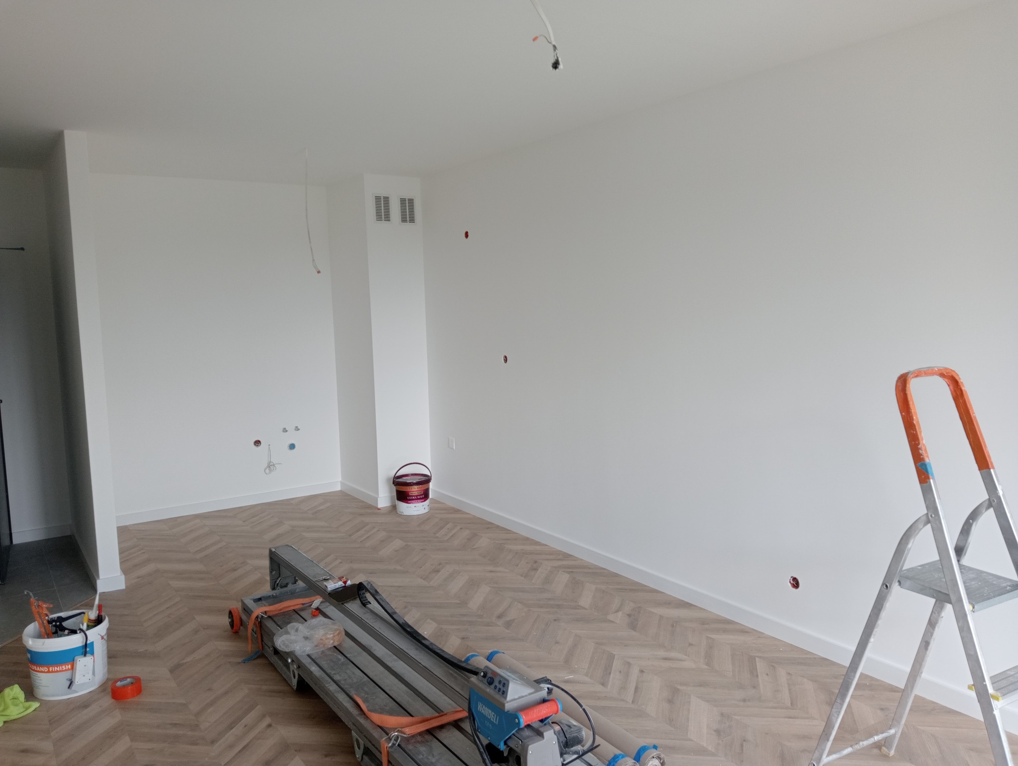 Białe, odświeżone wnętrze z parkietem w jodełkę, widoczna drabina malarska, puszka z farbą i narzędzia malarskie, przygotowane do dalszych prac wykończeniowych.