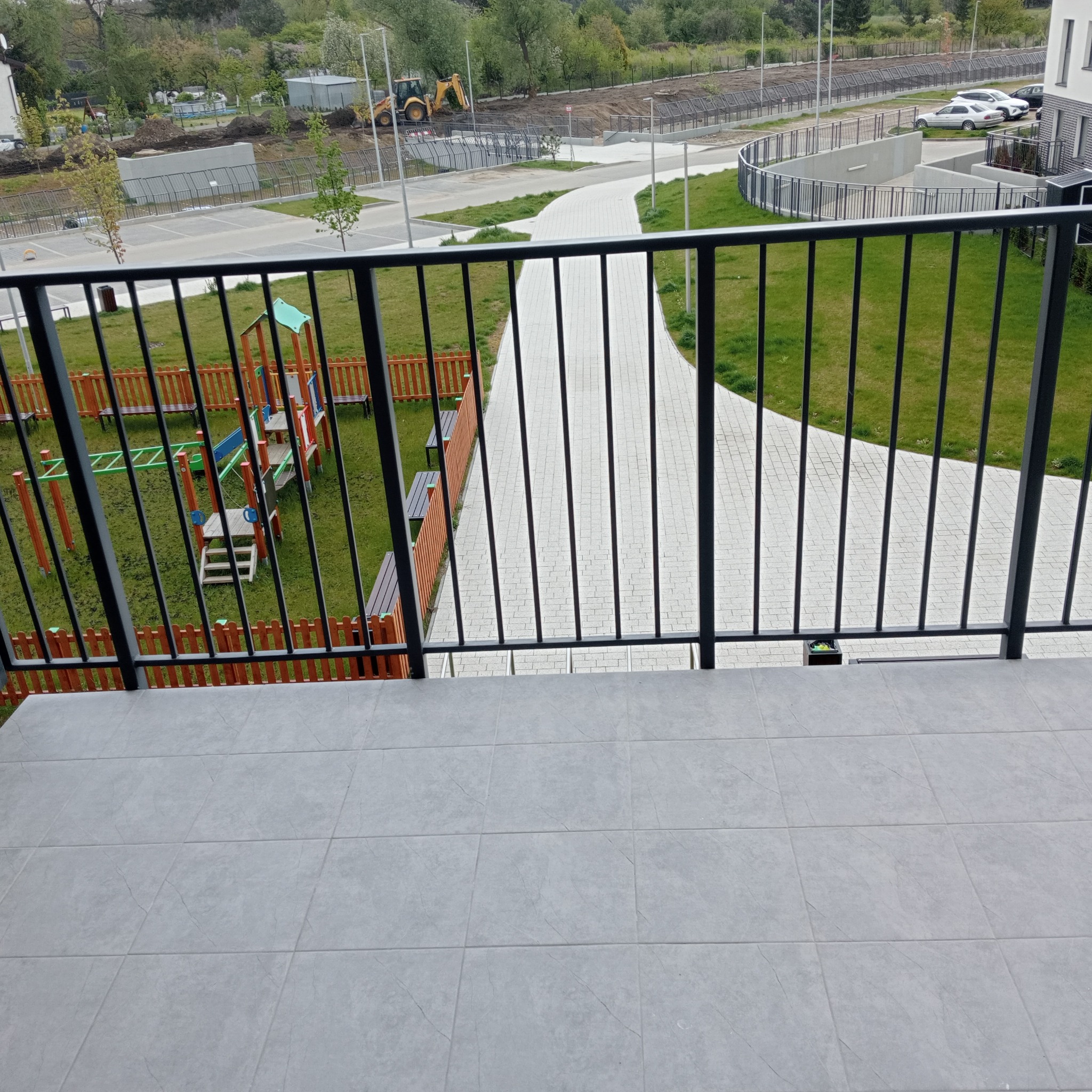 Widok z balkonu na plac zabaw, chodnik z kostki i teren budowy z koparką w tle, przez czarne metalowe barierki.