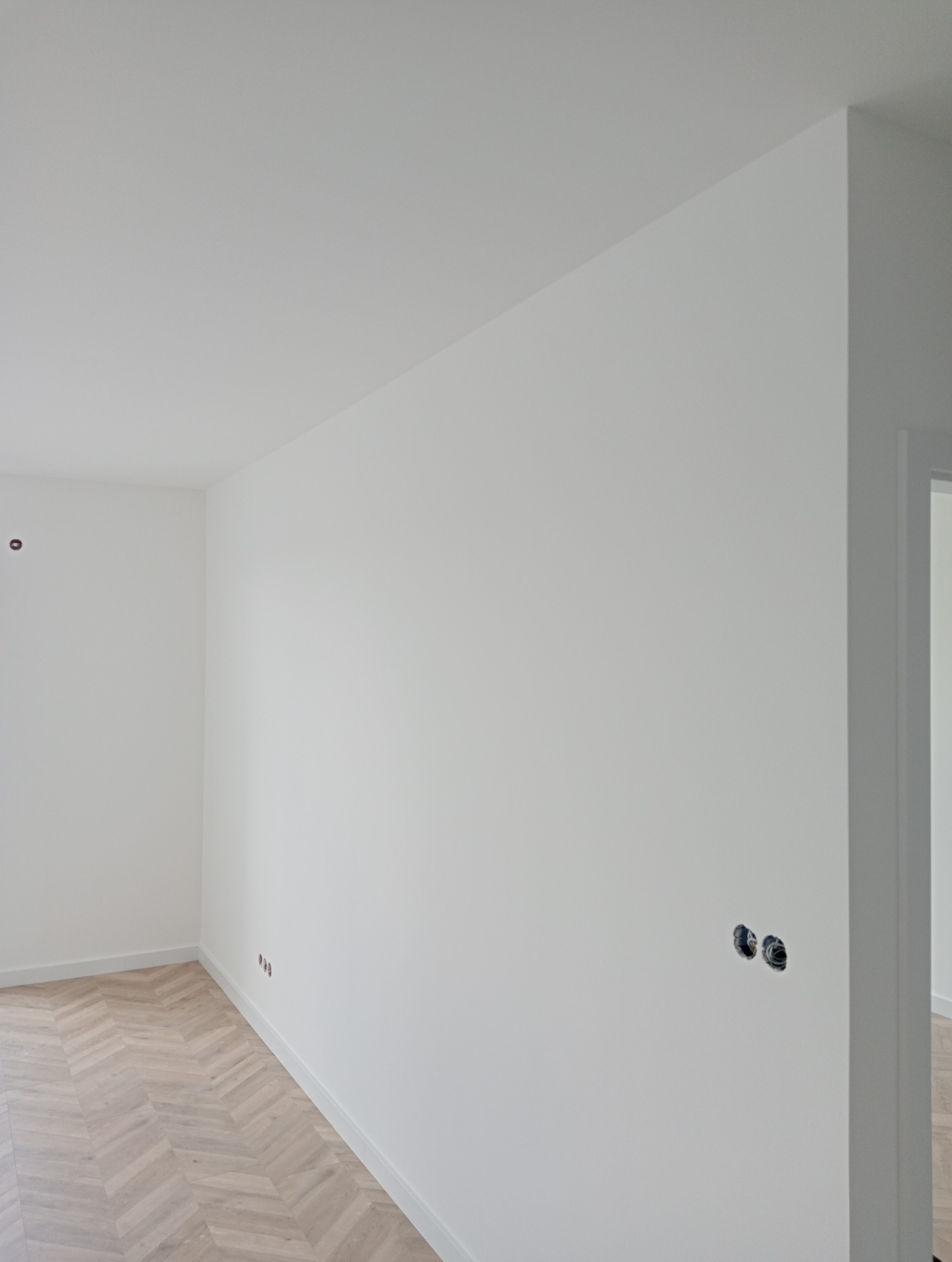 Puste, białe wnętrze z parkietem w jodełkę, widoczne puszki elektryczne na ścianie przed malowaniem.