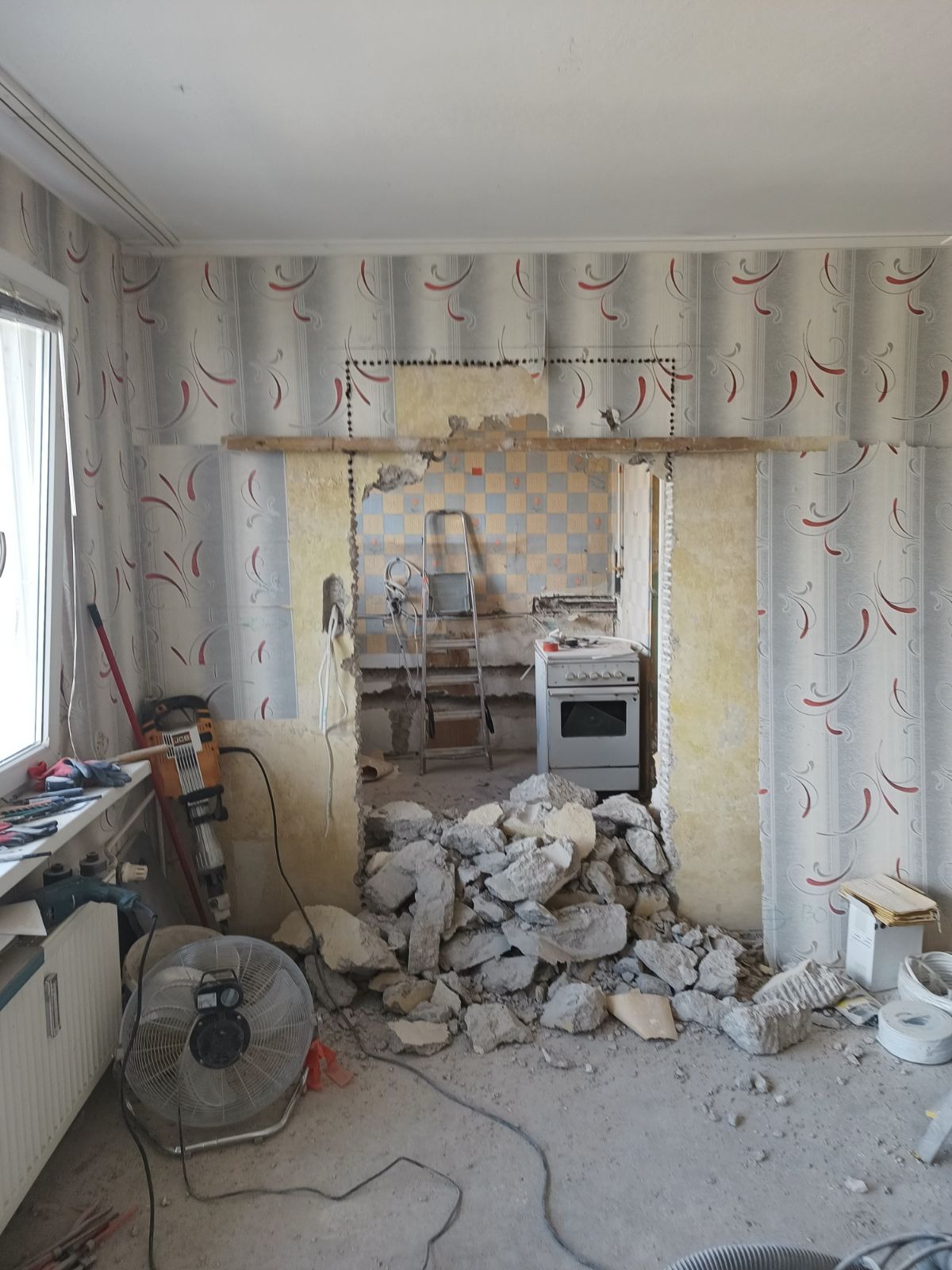 Demontaż ściany działowej między pokojem a kuchnią, widoczne gruz, odsłonięte instalacje, drabina i kuchenka w tle, ściany z tapetą w pokoju.