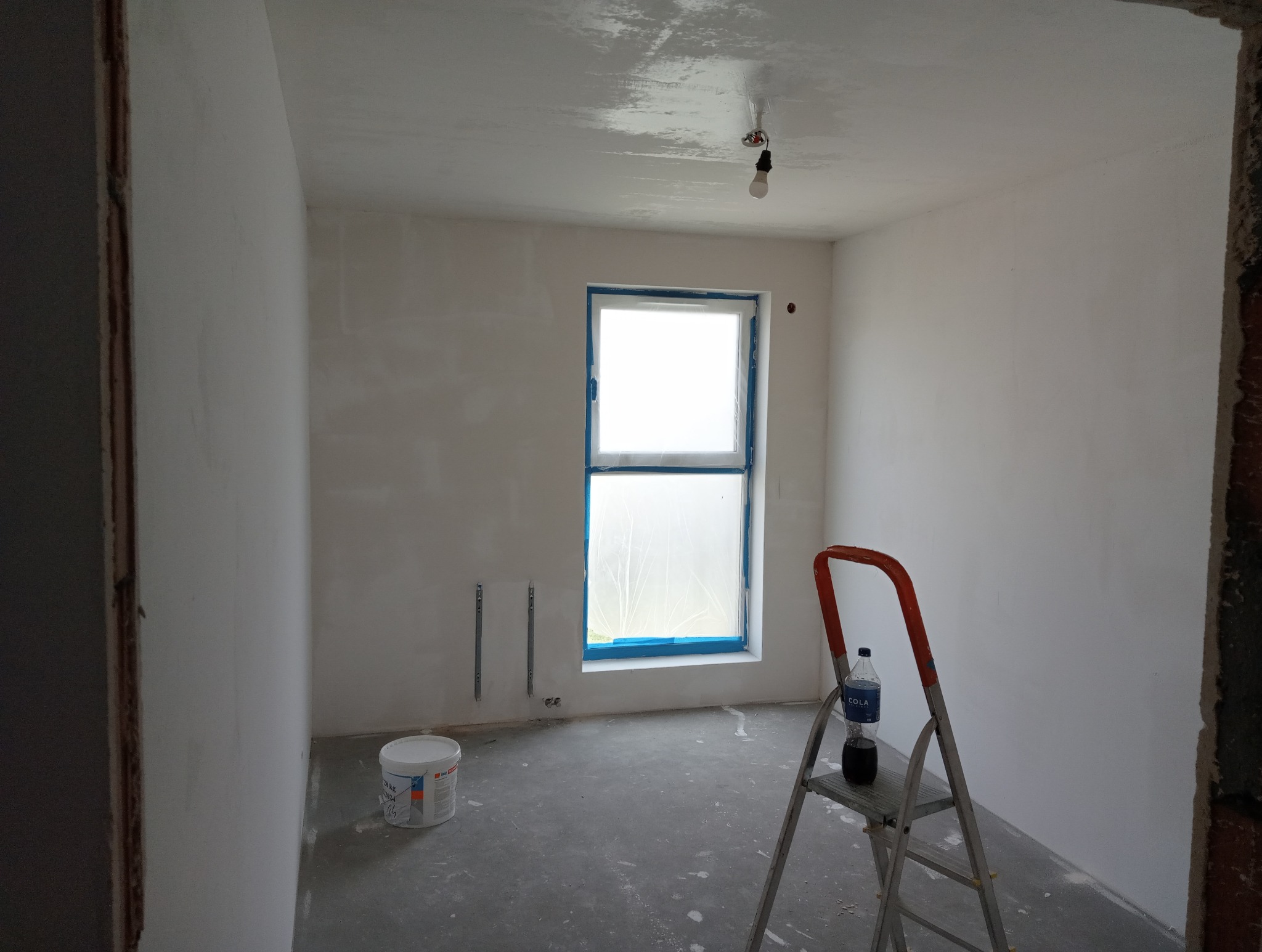 Pokój w trakcie prac wykończeniowych: świeżo zagruntowany sufit, zabezpieczone okno folią, drabina z butelką Coli, wiadro farby przy ścianie.