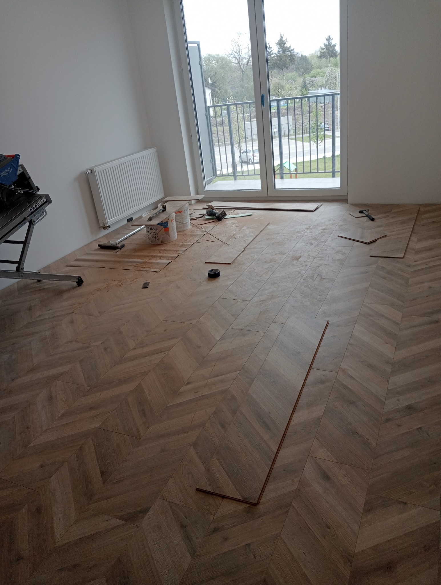 Układanie parkietu w jodełkę w jasnym pomieszczeniu z dużym oknem balkonowym, widoczne narzędzia i materiały budowlane podczas pracy.