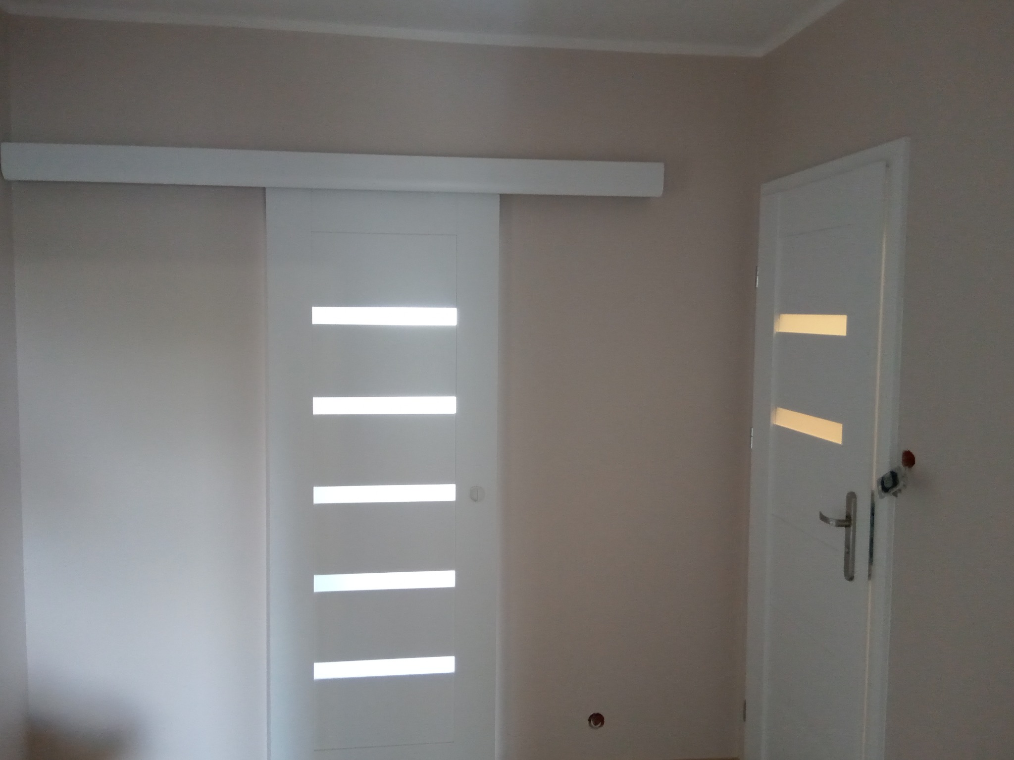 Białe, przesuwne drzwi z poziomymi przeszkleniami, zamontowane w jasnym wnętrzu. Widoczna również standardowa biała ościeżnica z klamką.