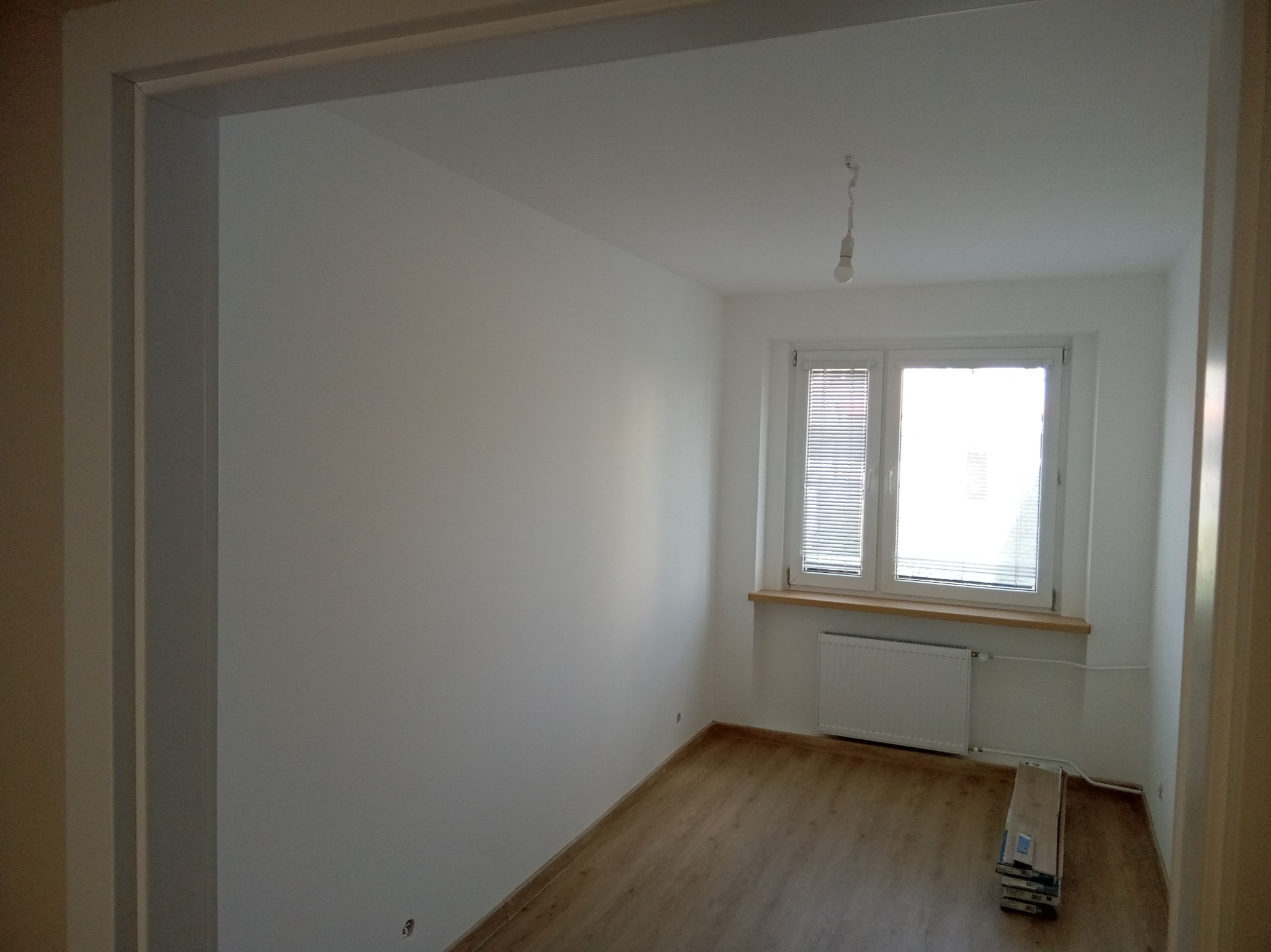 Jasne, pomalowane na biało wnętrze pokoju z drewnianą podłogą, widoczne przez framugę drzwi. Pod oknem grzejnik i stos paneli podłogowych.