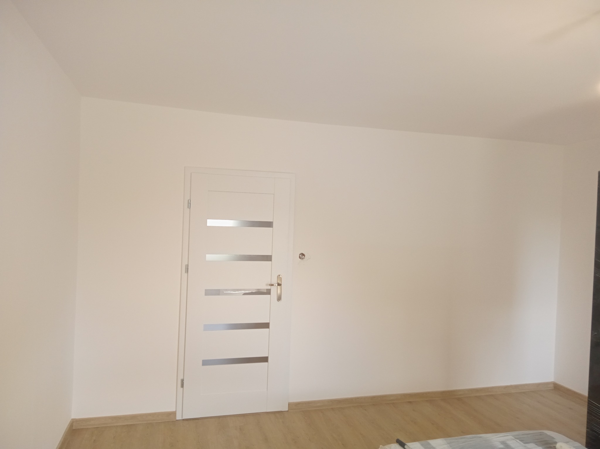 Białe drzwi z poziomymi, matowymi paskami w jasnym pokoju po remoncie. Widoczne świeżo pomalowane ściany i drewnopodobna podłoga. Minimalistyczny design.
